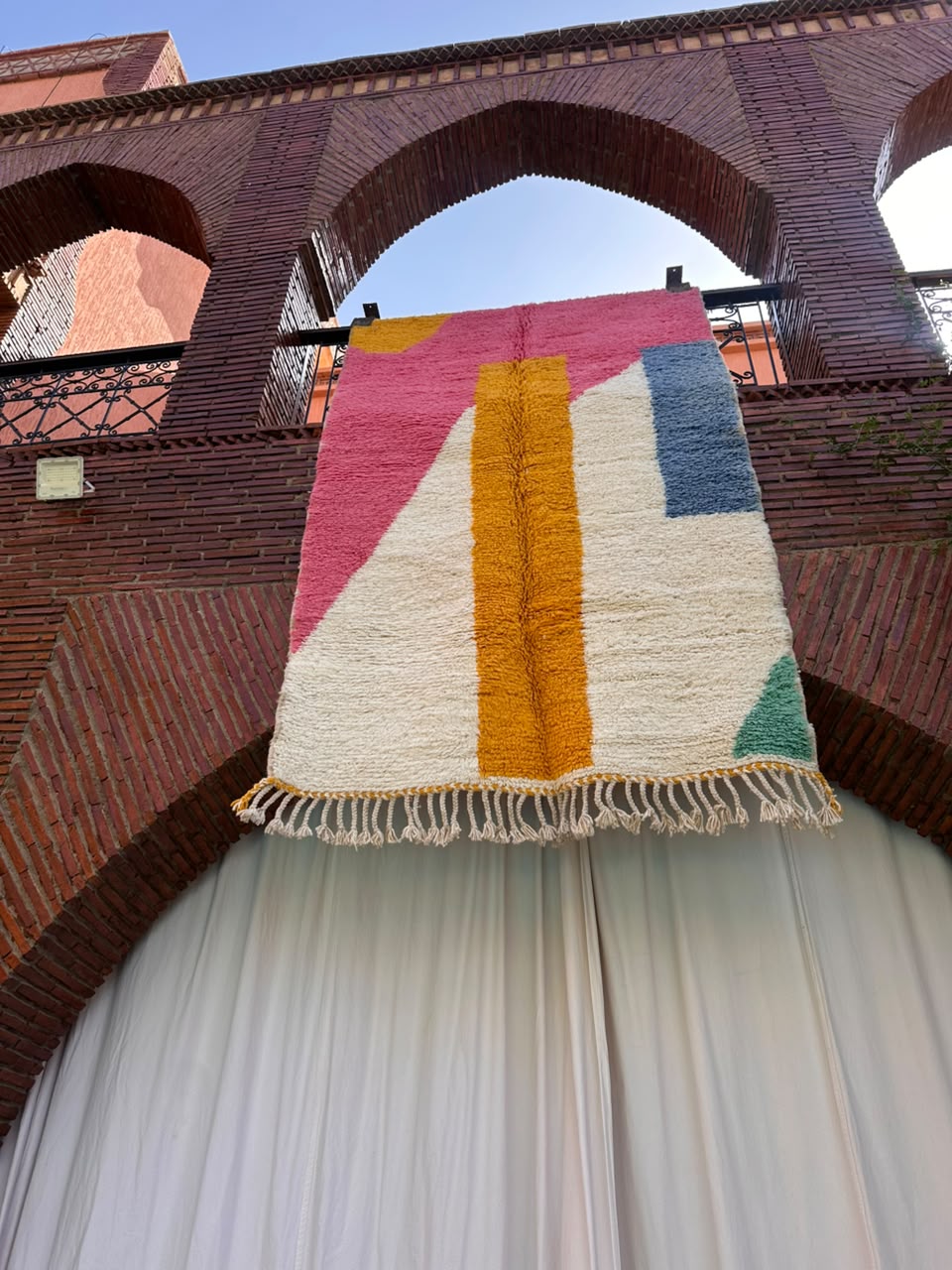 A colorful geometric area rug with cream, pink, mustard yellow, blue, and green blocks hangs from a black railing in front of a brick archway and white draped curtains.
