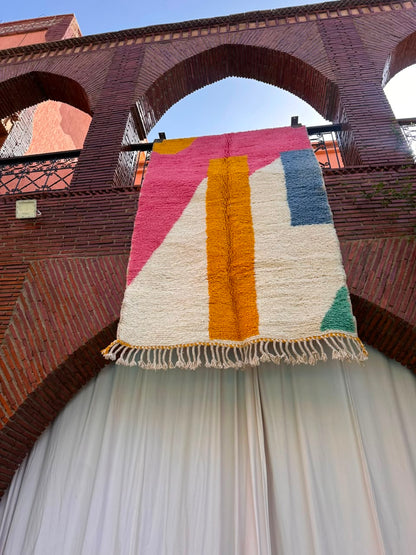 A colorful geometric area rug with cream, pink, mustard yellow, blue, and green blocks hangs from a black railing in front of a brick archway and white draped curtains.