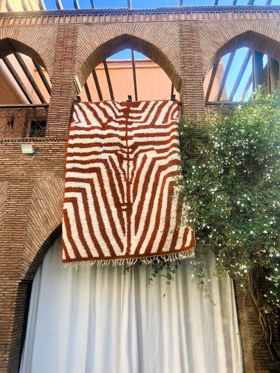 A large woven rug with a brown and cream wavy geometric pattern hangs from a black metal frame against a brick building facade with arched windows and green foliage.