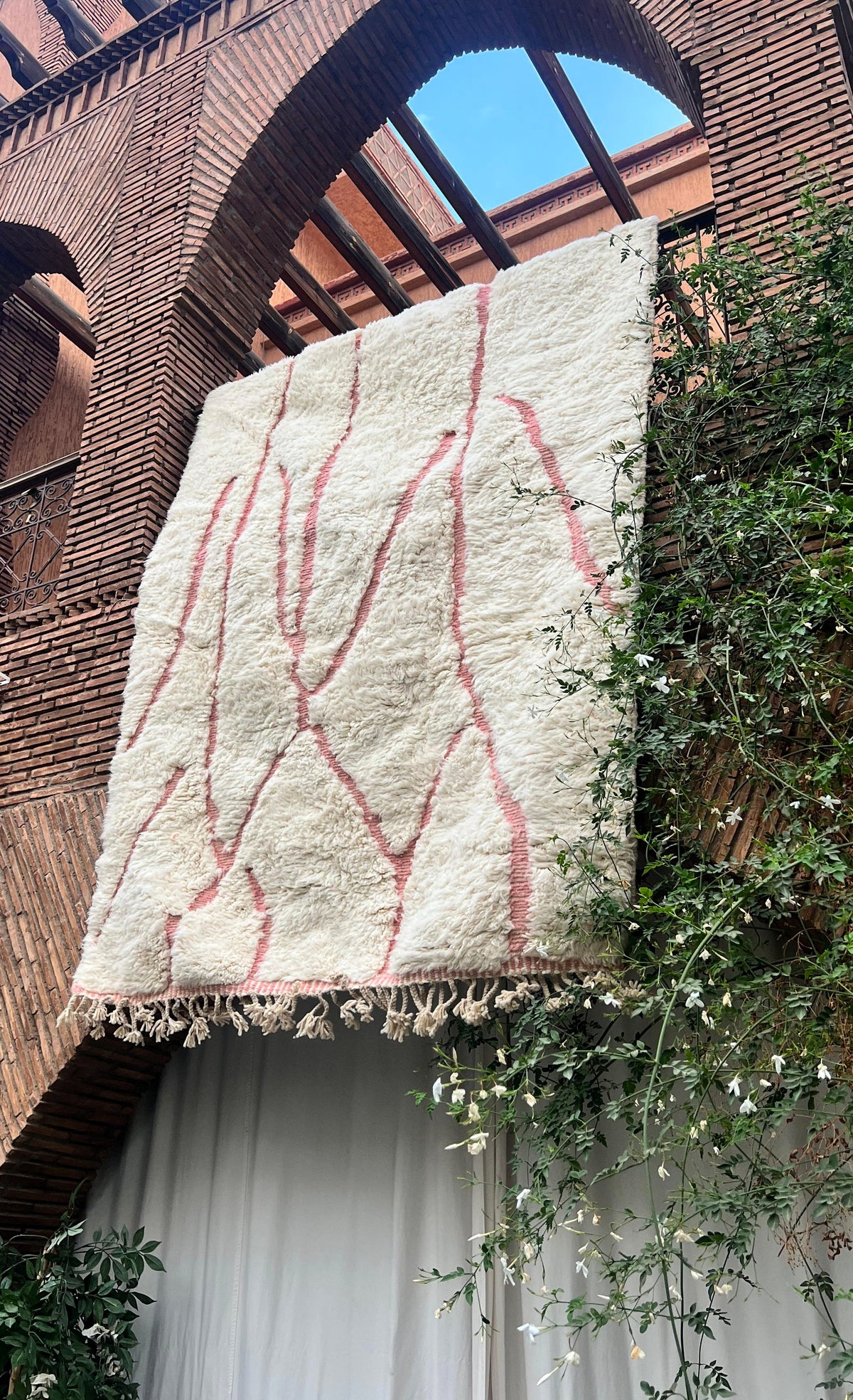 A large off-white wool rug with a pink abstract line pattern and tasseled fringe, hanging against a brick building wall with green vines and a white curtain below.