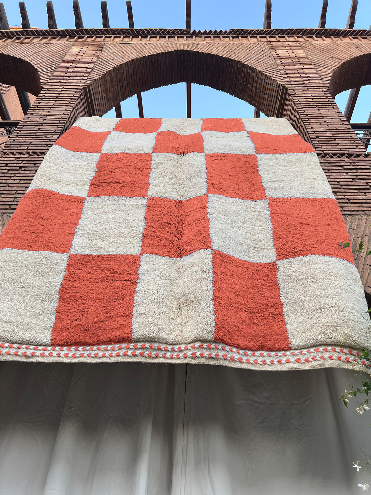 A large checkered wool rug with alternating rust orange and cream squares, draped horizontally against a brick archway structure.
