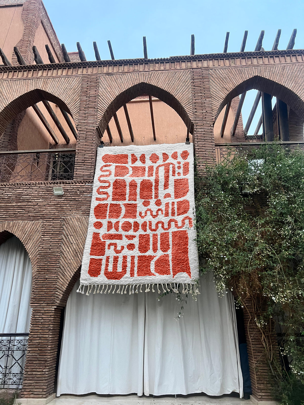 A large woven tapestry with red geometric patterns on a white background hangs from a brick building with arched windows and white curtains below.