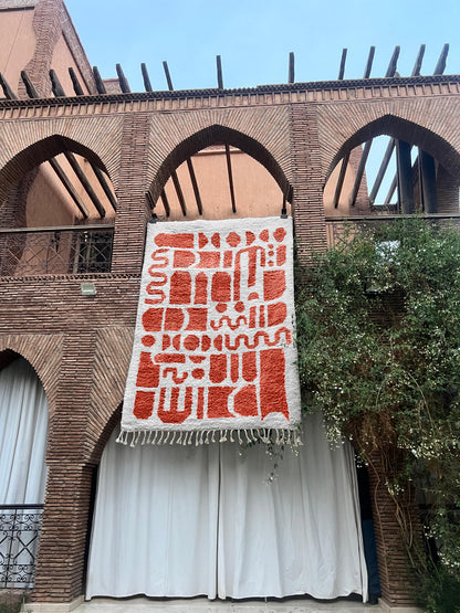 A large woven tapestry with red geometric patterns on a white background hangs from a brick building with arched windows and white curtains below.