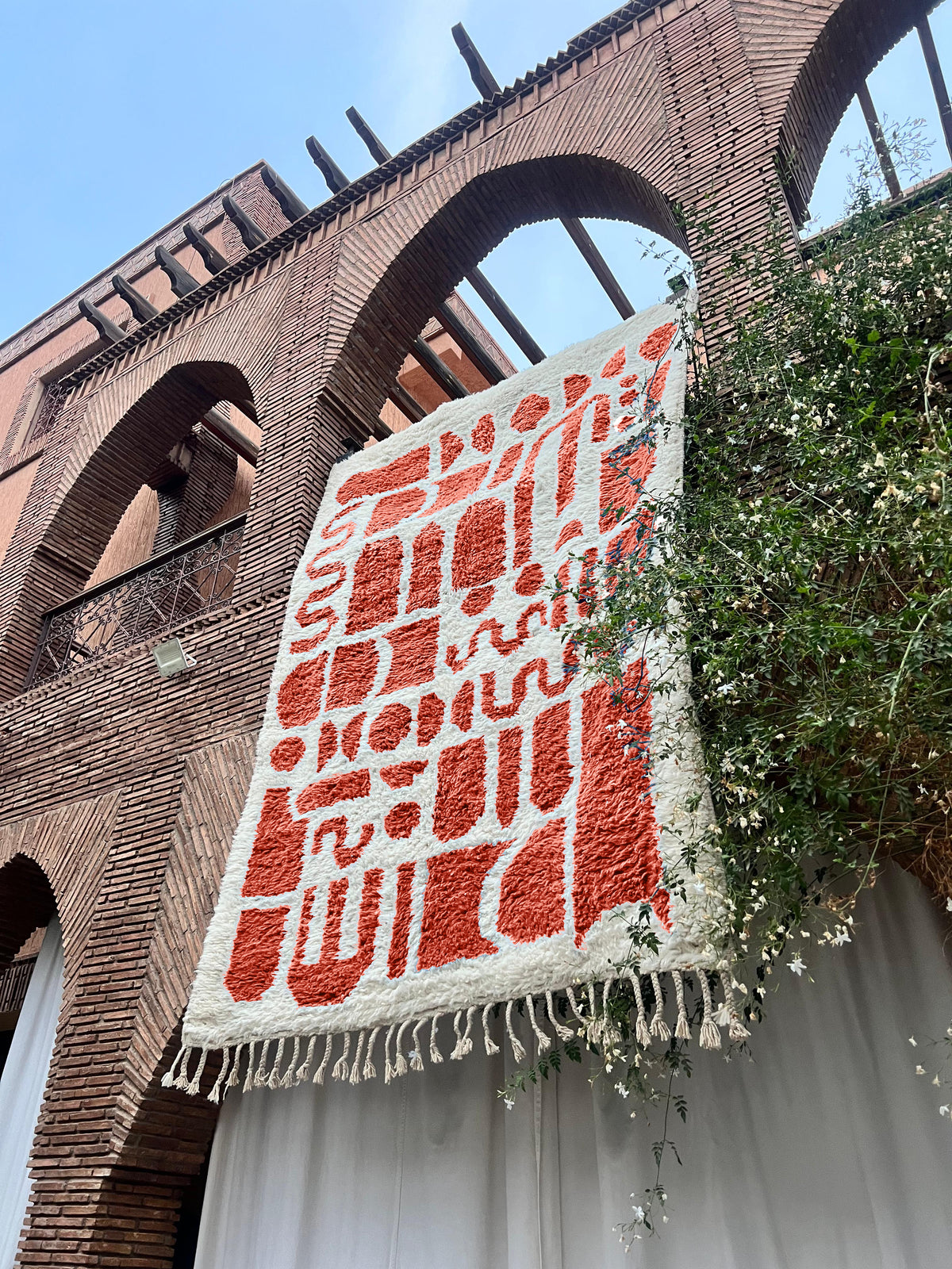 A large woven textile with cream and terracotta abstract patterning and fringe, hanging against a brick building facade with greenery.