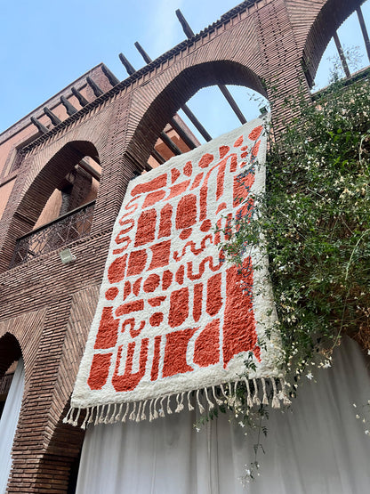 A large woven textile with cream and terracotta abstract patterning and fringe, hanging against a brick building facade with greenery.