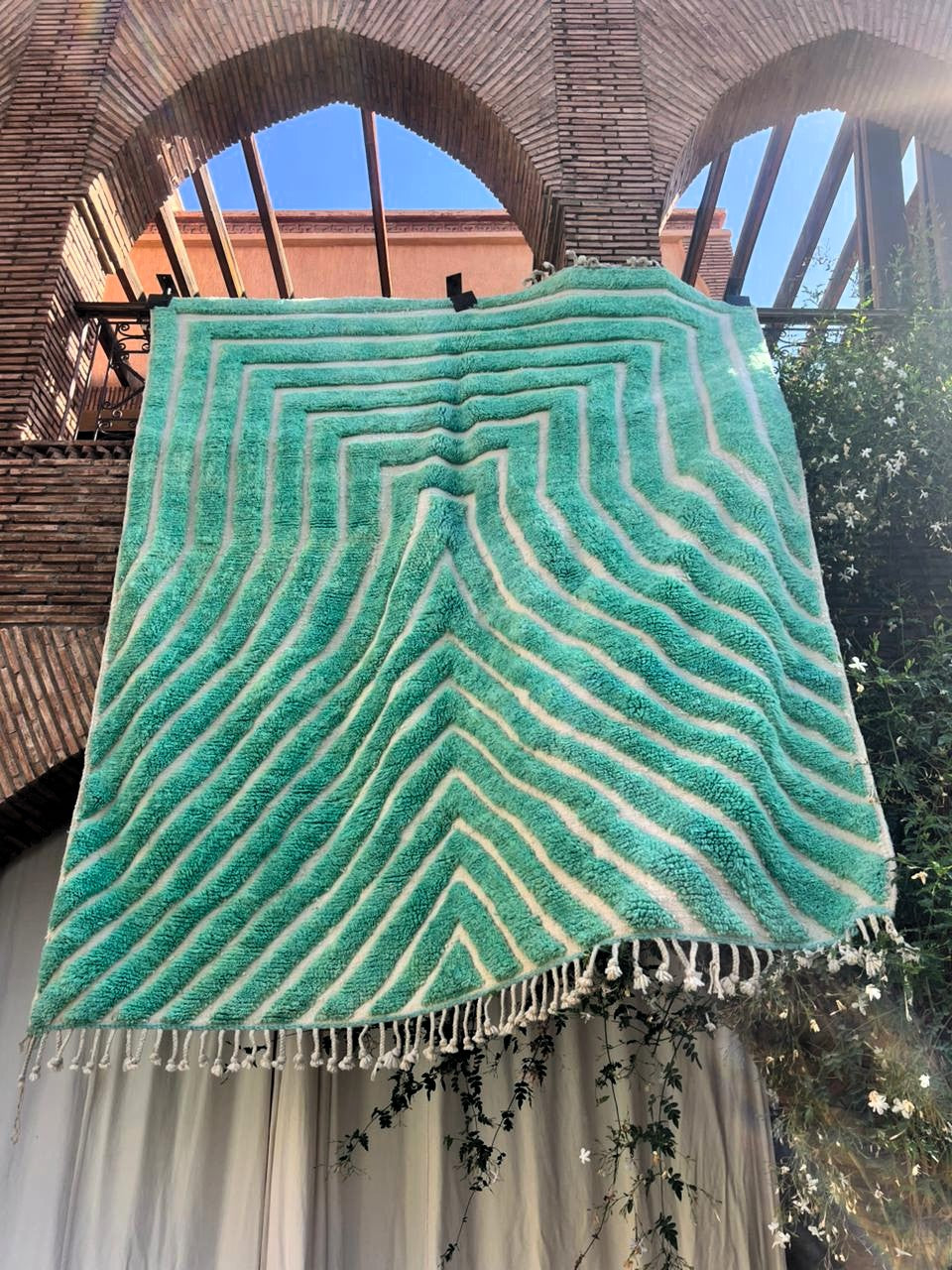 A large rectangular turquoise and cream striped tigmee moroccan rug with fringe, hanging outdoors under a brick archway structure.
