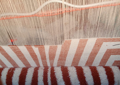 Close-up of a woven textile with alternating wide and narrow vertical stripes in rust red and off-white, with additional white and rust red horizontal bands and fine thread lines visible above.